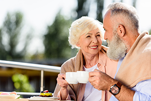 Elderly couple sitting outside smiling at each while toasting with coffee cups